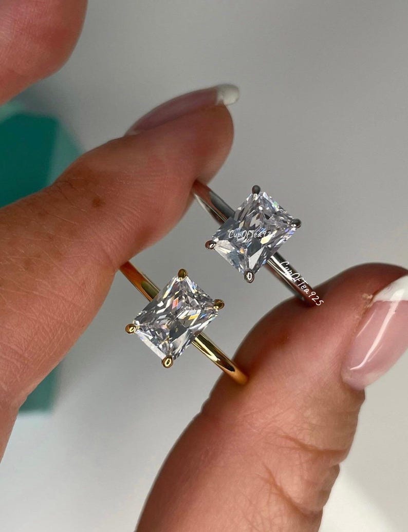 May include: Two engagement rings with square-shaped cubic zirconia stones. The ring on the left has a gold band, and the ring on the right has a silver band.