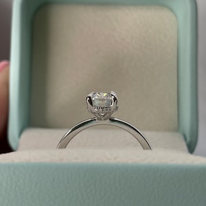 May include: A silver engagement ring with a round, clear diamond in a prong setting. The ring is in a light blue ring box.
