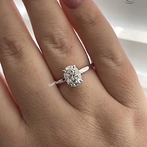 May include: A silver ring with an oval-shaped diamond. The ring is on a person's finger.