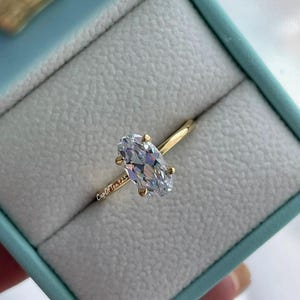Puede incluir: Un anillo de oro con una gran piedra preciosa ovalada transparente. El anillo est&aacute; en una caja de anillo azul claro. El texto "Cup Of Tea 925" es visible en la banda del anillo.