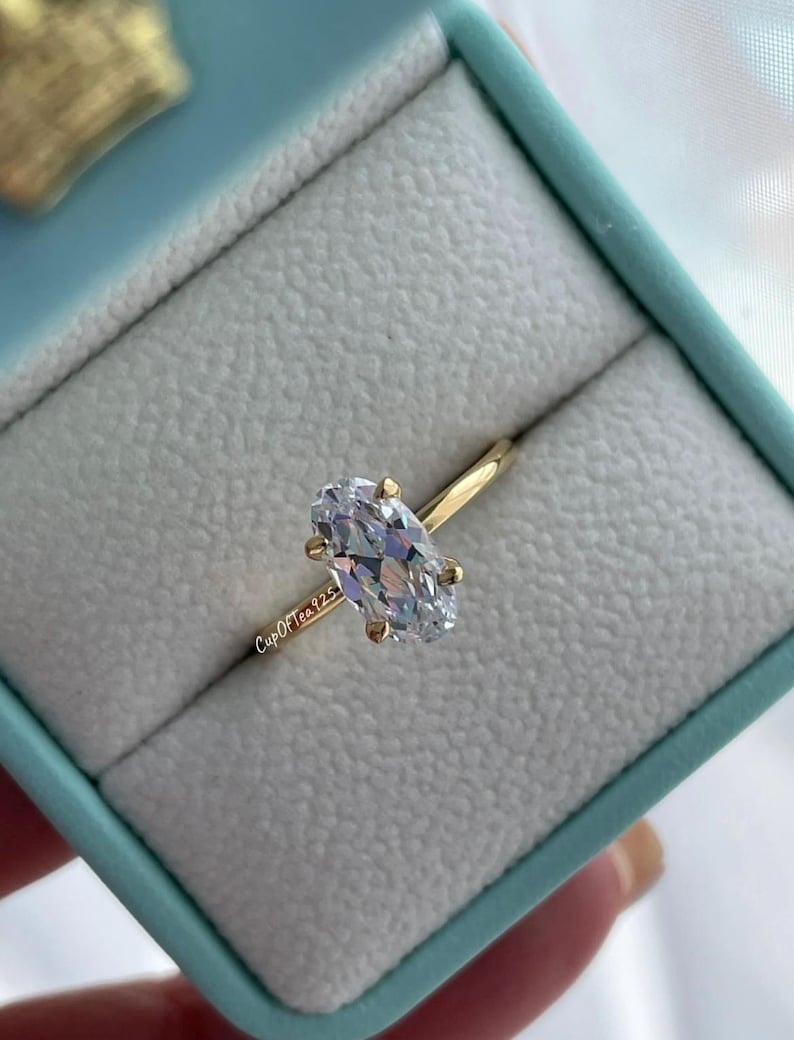 Puede incluir: Un anillo de oro con una gran piedra preciosa ovalada transparente. El anillo est&aacute; en una caja de anillo azul claro. El texto "Cup Of Tea 925" es visible en la banda del anillo.