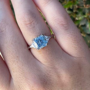 May include: A silver ring with a large, square-shaped, light blue gemstone surrounded by two smaller, triangular-shaped, clear gemstones.