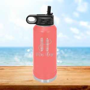 May include: A coral pink stainless steel water bottle with a black lid and a black carrying handle. The bottle has a silver base and is engraved with two pencils forming the letter H and the text "Mrs. Haze".