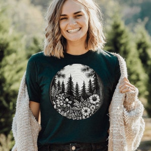 May include: A dark green t-shirt with a black and white illustration of a forest scene. The illustration is circular and features trees, flowers, and ferns.