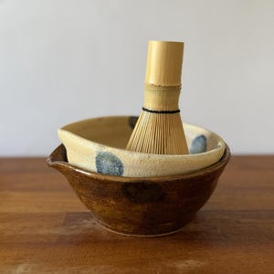 May include: A bamboo matcha whisk rests inside a ceramic bowl. The bowl features a two-tone design with a dark brown glazed lower section and a lighter, speckled cream-coloured upper rim decorated with small, circular blue painted accents.