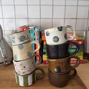 May include: Two stacks of four ceramic mugs each. The left stack features a floral pattern, blue dots, pink and gold dots, and green stripes. The right stack includes a white mug with black dots, a solid black mug, a brown mug with dots, and a solid brown mug.