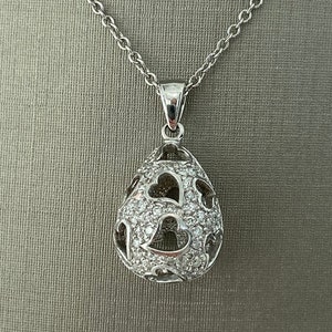 Könnte beinhalten: Eine silberne Kettenkette mit einem Anhänger in Tropfenform. Der Anhänger ist mit kleinen, klaren Steinen besetzt und hat ein ausgeschnittenes Design mit Herzen.