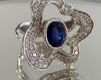 May include: A silver ring with a floral design, featuring a large oval-cut, deep blue gemstone at the center. The ring is encrusted with small, clear gemstones, and has a polished, reflective surface.