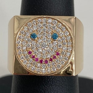 Puede incluir: Un anillo de oro con un engaste cuadrado que presenta un diseño de cara sonriente. La cara sonriente está hecha de pequeños diamantes blancos con dos piedras azules para los ojos y una fila de piedras rosas para la boca.