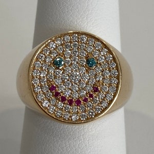 Puede incluir: Un anillo de oro con un diseño de cara sonriente. La cara sonriente está hecha de pequeños diamantes blancos y tiene dos piedras preciosas azules para los ojos y una fila de piedras preciosas rosas para la boca.