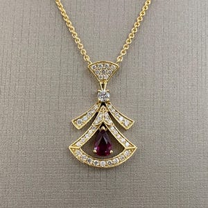 Könnte beinhalten: Eine goldene Halskette mit einem Anhänger, der einen birnenförmigen Rubin zeigt, der von einem Heiligenschein aus Diamanten umgeben ist. Der Anhänger ist mit einem geometrischen Muster verziert und an einer zarten Kette befestigt.