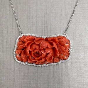 May include: A delicate necklace with a carved coral pendant. The pendant features a floral design with intricate details and is surrounded by a border of sparkling diamonds.