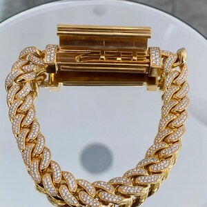 A gold chain bracelet with a hinged clasp. The chain is made of interlocking links, each of which is encrusted with small, clear stones. The clasp is also encrusted with stones.