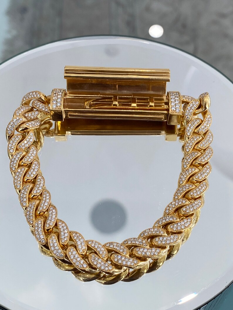 A gold chain bracelet with a hinged clasp. The chain is made of interlocking links, each of which is encrusted with small, clear stones. The clasp is also encrusted with stones.