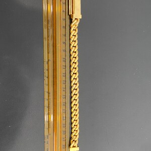 A gold chain bracelet with a clasp on each end. The chain is a curb style and is measured against a ruler that shows the bracelet is 21 centimeters long.