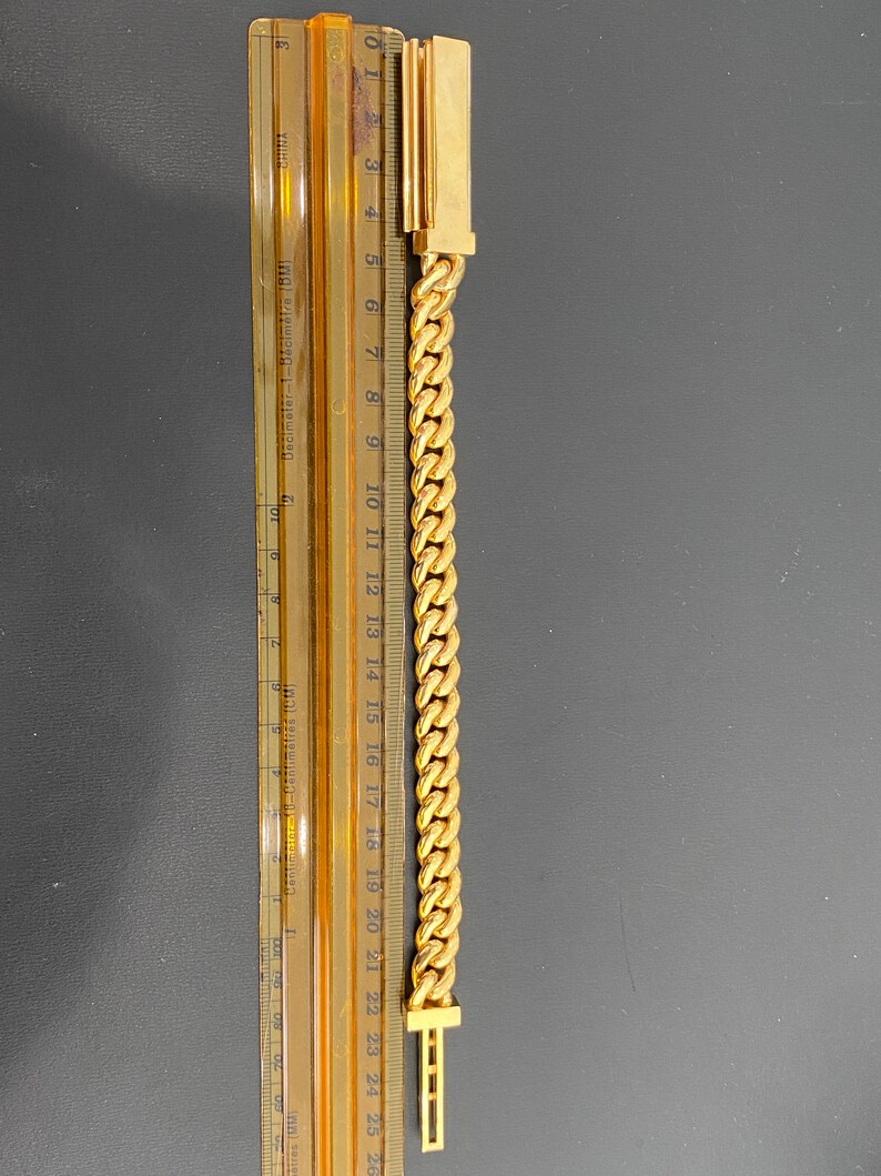 A gold chain bracelet with a clasp on each end. The chain is a curb style and is measured against a ruler that shows the bracelet is 21 centimeters long.