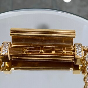 A gold chain bracelet with a diamond-encrusted clasp. The clasp is open, revealing a gold interior with a series of small, rectangular compartments.