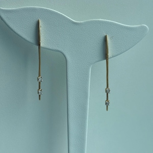 Könnte beinhalten: Ein Paar goldene Ohrringe mit jeweils drei kleinen Diamanten. Die Ohrringe befinden sich auf einem weißen Präsentationsständer.