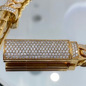 A gold chain bracelet with a clasp that is covered in small, white diamonds. The bracelet is made of thick, interlocking links. The clasp is a rectangular shape and is also covered in diamonds.