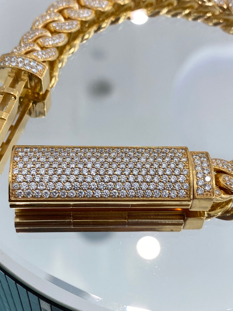 A gold chain bracelet with a clasp that is covered in small, white diamonds. The bracelet is made of thick, interlocking links. The clasp is a rectangular shape and is also covered in diamonds.