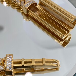 A gold bracelet with a diamond-encrusted clasp. The clasp is open, revealing the inner workings of the clasp. The bracelet is on a white surface.