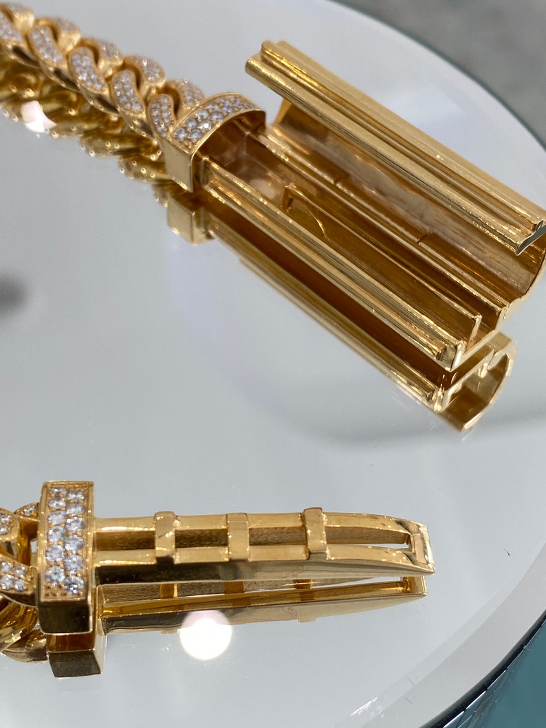 A gold bracelet with a diamond-encrusted clasp. The clasp is open, revealing the inner workings of the clasp. The bracelet is on a white surface.