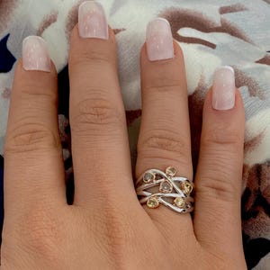 Peut inclure: Une bague en argent et or avec un design délicat et entrelacé. La bague est sertie de plusieurs petites pierres précieuses facettées dans des tons de jaune et de brun.
