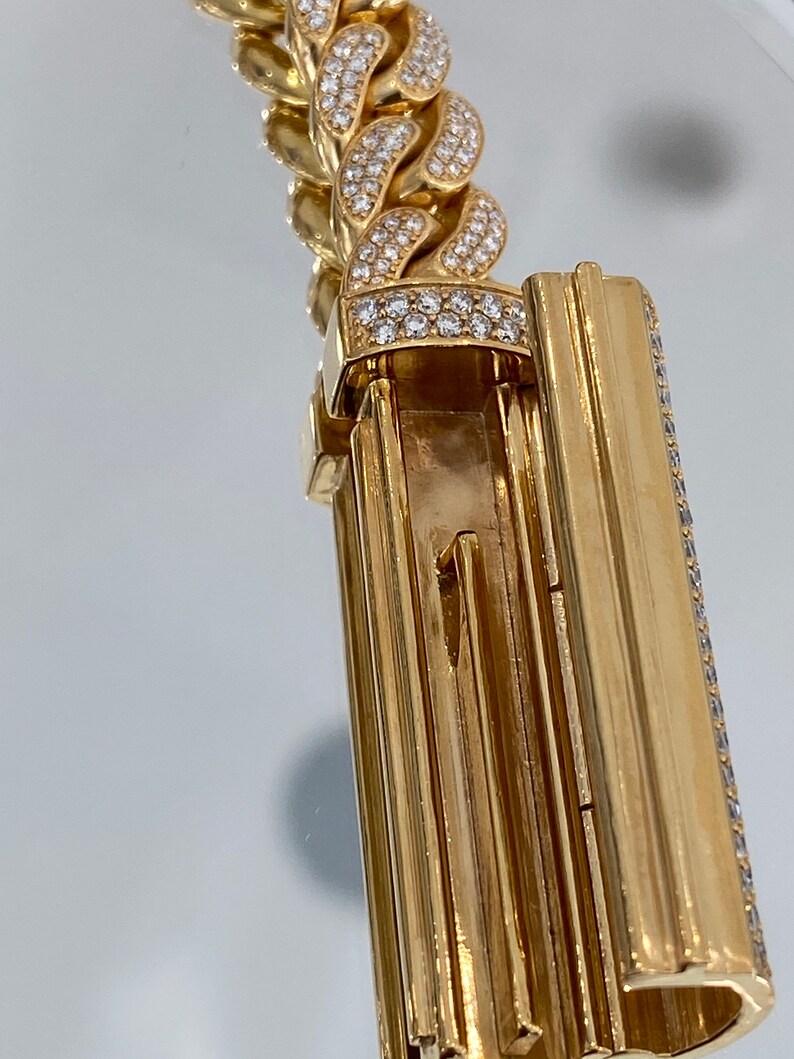 A gold chain bracelet with a clasp. The bracelet is covered in small, clear diamonds. The clasp is a hinged design with a series of vertical lines.