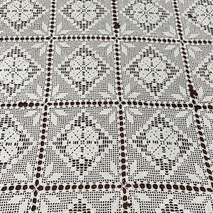 May include: Cream-colored, crocheted tablecloth with a repeating square pattern. Each square features a floral design in the center, surrounded by intricate openwork. The tablecloth is made of a soft, textured yarn.