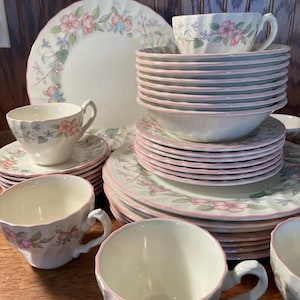 May include: A collection of floral-patterned china dishes, including plates, bowls, and teacups. The set features a white base with pink floral designs and pink trim. The arrangement includes stacked plates and bowls, with teacups and saucers.