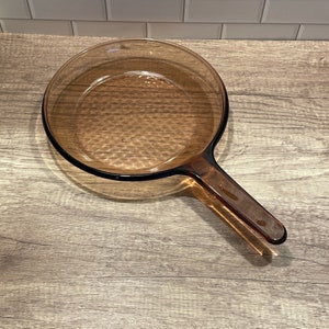 May include: A clear, amber-colored glass skillet with a long handle. The skillet has a round cooking surface and a black rim. The handle is angled and extends from the side of the pan. The skillet is sitting on a wooden surface.
