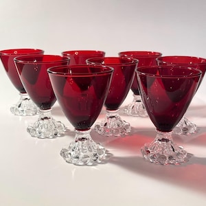 May include: A set of seven ruby red glass goblets with clear, textured, hobnail-patterned bases. Each goblet features a deep red, smooth, conical bowl supported by a short, thick stem and a wide, circular foot decorated with raised glass beads.