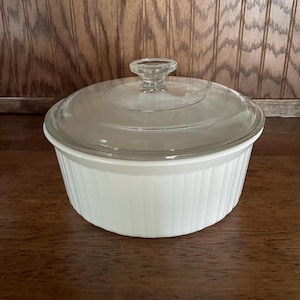 May include: A white, ribbed casserole dish with a clear glass lid. The lid has a small, round handle. The dish is sitting on a dark wooden surface. The dish is designed for cooking and serving.