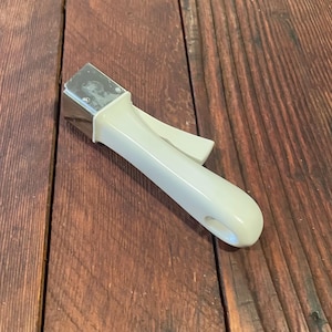 May include: Cream-colored pot handle with a metal attachment. The handle has a curved design with a small lever and a hole at the end. The handle is resting on a wooden surface.