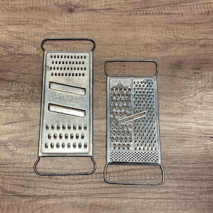 May include: Two metal cheese graters. The grater on the left is labeled "Bromco" and has a rectangular shape with a handle on each end. The grater on the right is labeled "All-Purpose" and has a rectangular shape with a handle on each end.