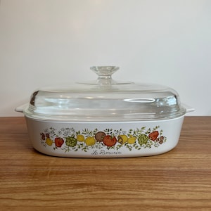May include: White rectangular casserole dish with a clear glass lid. The dish features a colorful border of fruits and vegetables, including tomatoes, lemons, and artichokes. The words "Le Romarin" are printed on the front.