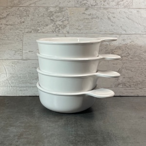May include: A stack of four white ceramic soup bowls with handles. The bowls are round with a slightly flared rim and a small handle on one side. The bowls are stacked on top of each other.