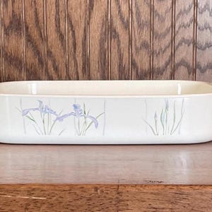 May include: A white rectangular baking dish with a floral design featuring blue irises and green leaves. The dish has two handles on the sides.