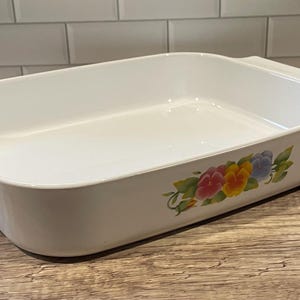 May include: White rectangular baking dish with handles. The dish features a floral design with pink, yellow, and blue flowers. The dish is on a wooden surface.