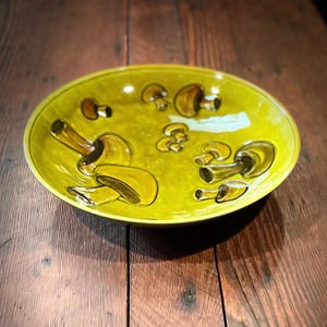 May include: A round, yellow ceramic serving dish with a hand-painted mushroom design. The mushrooms are depicted in shades of brown and yellow, with a glossy finish. The dish is set on a wooden surface.