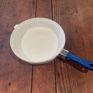 May include: A white enamel pan with a blue handle. The pan is round with a wide rim and a single handle. The handle is blue with a metal attachment. The pan is sitting on a wooden surface.