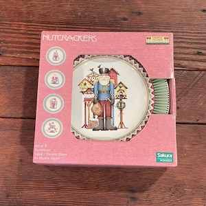 May include: A set of four stoneware salad/dessert plates in a pink box. The plates feature a detailed illustration of a nutcracker figure with birdhouses. The box reads "NUTCRACKERS" and "DEBBIE MUMM".