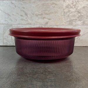 May include: A round, ribbed, purple container with a matching maroon lid. The container is made of a translucent material, and the lid sits snugly on top. The container is designed for food storage.