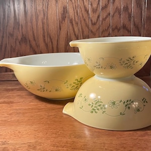 May include: Three yellow Pyrex mixing bowls with a floral design. The bowls are stacked and arranged on a wooden surface. The bowls have a light yellow exterior and a white interior. The floral design is green.