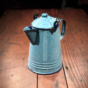 May include: A vintage-style, light blue enamelware coffee pot with a speckled pattern. The pot features a lid, handle, and spout, all in matching blue with dark blue trim. The pot is designed for hanging over a fire.