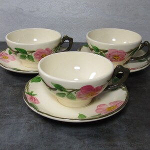 May include: Three white teacups with floral designs and matching saucers. The cups and saucers feature a pink rose pattern with green leaves.