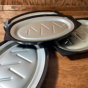 May include: A collection of oval-shaped, silver-colored metal trays with black rims. Each tray has a series of parallel, raised lines on the surface. The trays are stacked and arranged on a wooden surface.