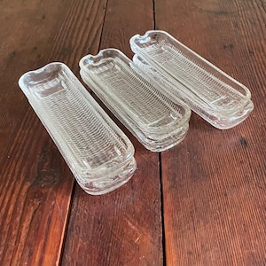 May include: A collection of clear glass corn-on-the-cob dishes. Each dish is rectangular with a curved end and a textured, woven pattern. The dishes are stacked in groups on a dark wood surface.