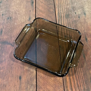 May include: A square, brown glass baking dish with handles. The dish is empty and sits on a wooden surface. The glass has a slight tint, and the edges are rounded.
