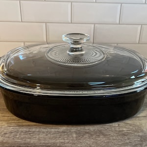 May include: An oval, black casserole dish with a clear glass lid. The lid has a ribbed design and a clear glass knob. The dish has two black handles on either side. The dish is sitting on a wooden surface.